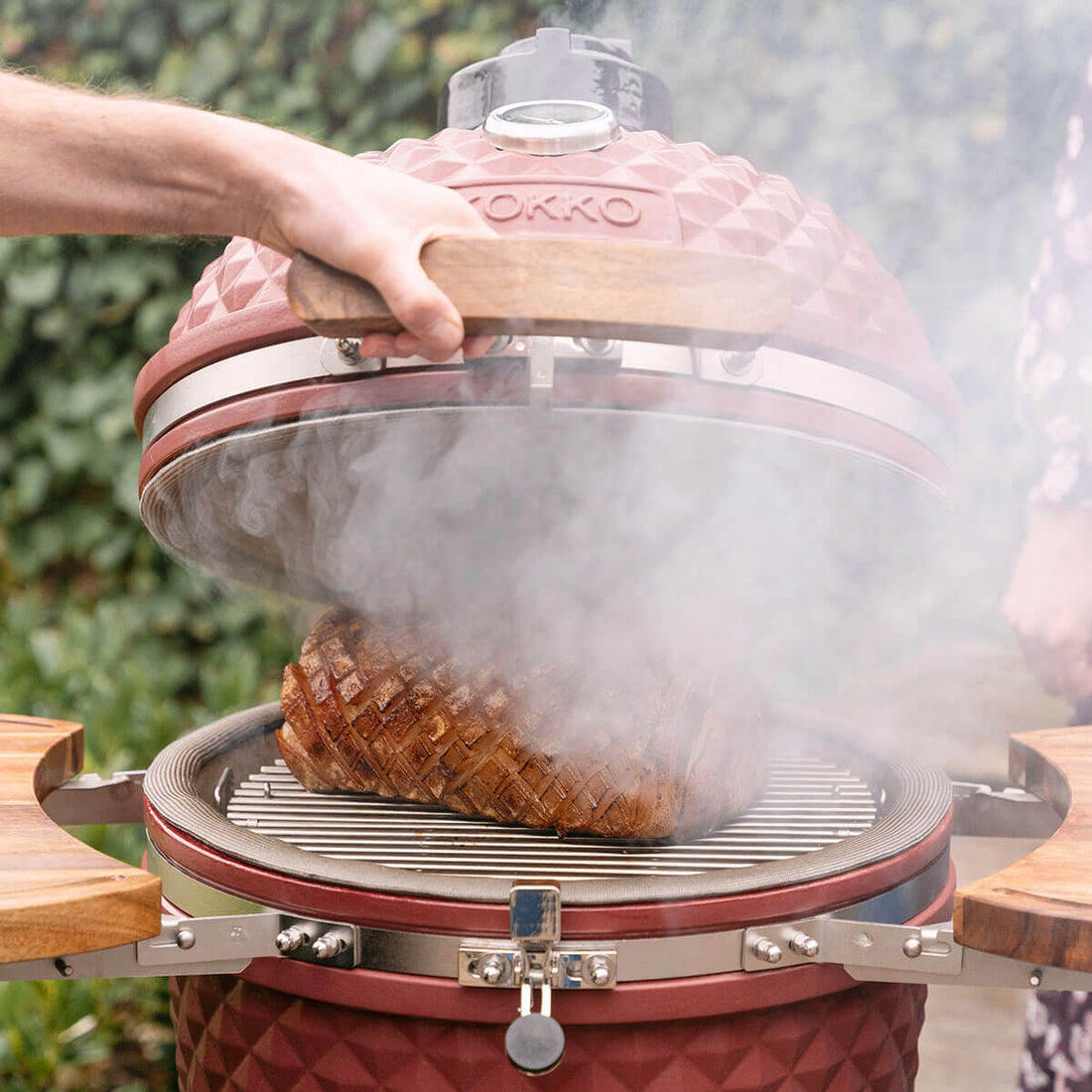 Uma mão levanta a tampa de um kamado Kokko vermelho óxido, revelando um pedaço de carne grelhada sobre a grelha em inox. Uma densa fumaça escapa da cuba, destacando a cozedura a lenha, a junta periférica e os tabuleiros laterais em madeira.
