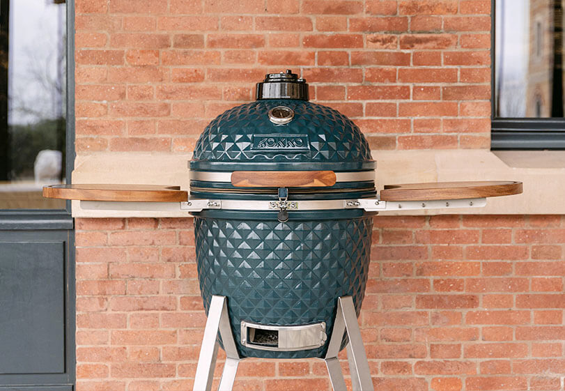 Kamado Kokko azul oceano colocado em suporte com prateleiras de madeira, visto de frente no exterior junto a uma parede de tijolos.