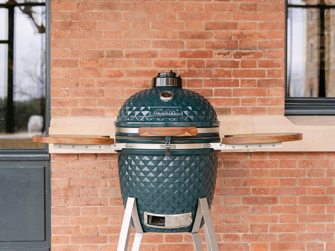 Kamado Kokko na cor verde oceano, instalado no exterior em frente a uma parede de tijolos. O barbecue está fechado, com tampa em cerâmica texturizada, termómetro integrado no topo, pega em madeira, fecho de segurança em inox e dois tabuleiros laterais em madeira de acácia. O conjunto assenta sobre um suporte metálico com bandeja de ventilação inferior visível.