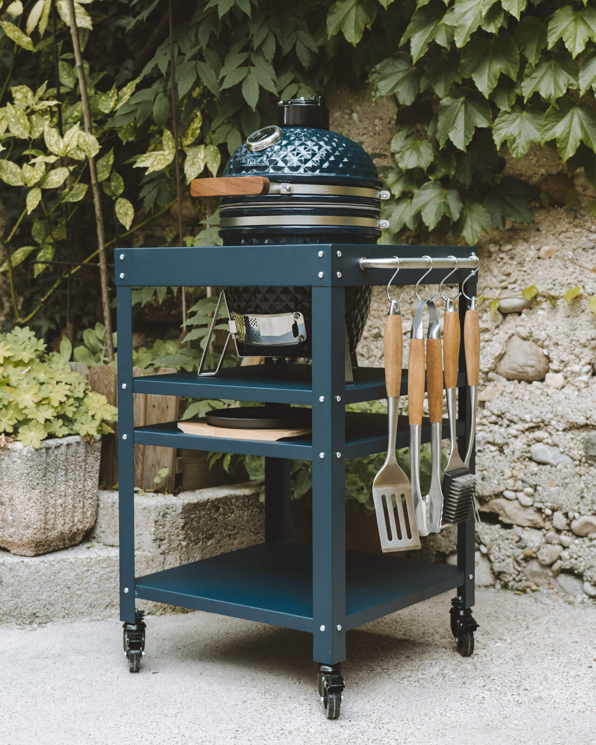 Kamado Kokko cor azul oceano colocado numa mesa de apoio metálica a condizer, com prateleiras de arrumação, recipiente para cinzas e utensílios pendurados na lateral.
