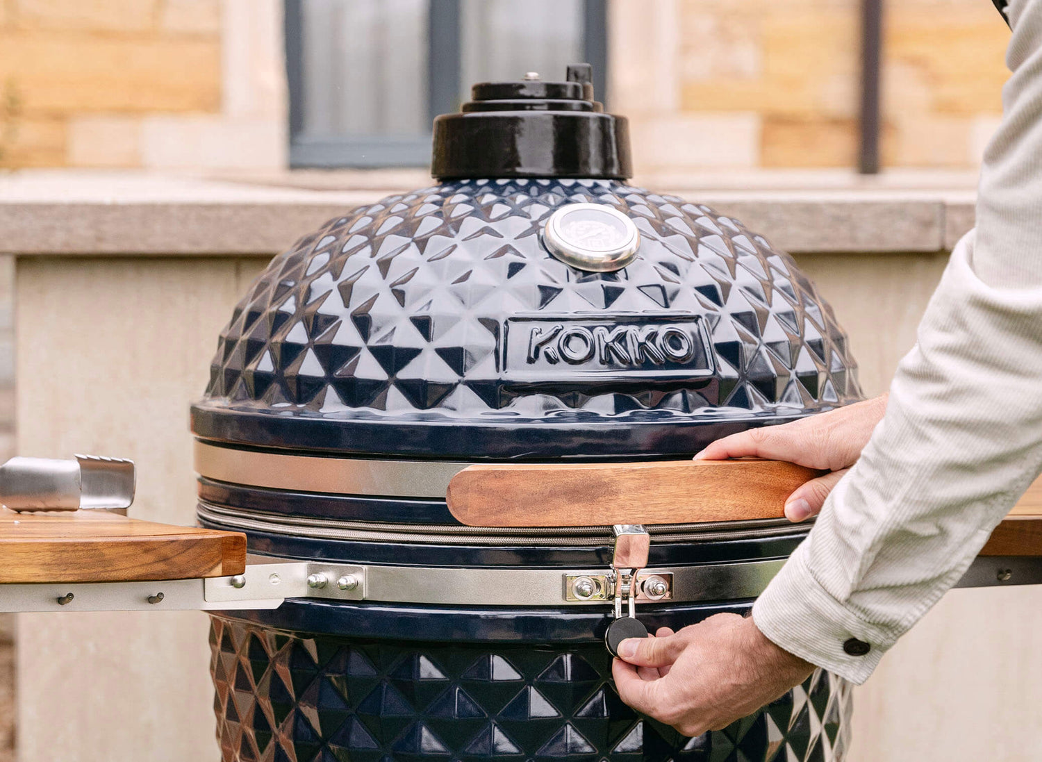 Mão a accionar o fecho de segurança de um kamado Kokko preto, com tampa em cerâmica texturada, pega em madeira e termómetro integrado, em utilização no exterior.