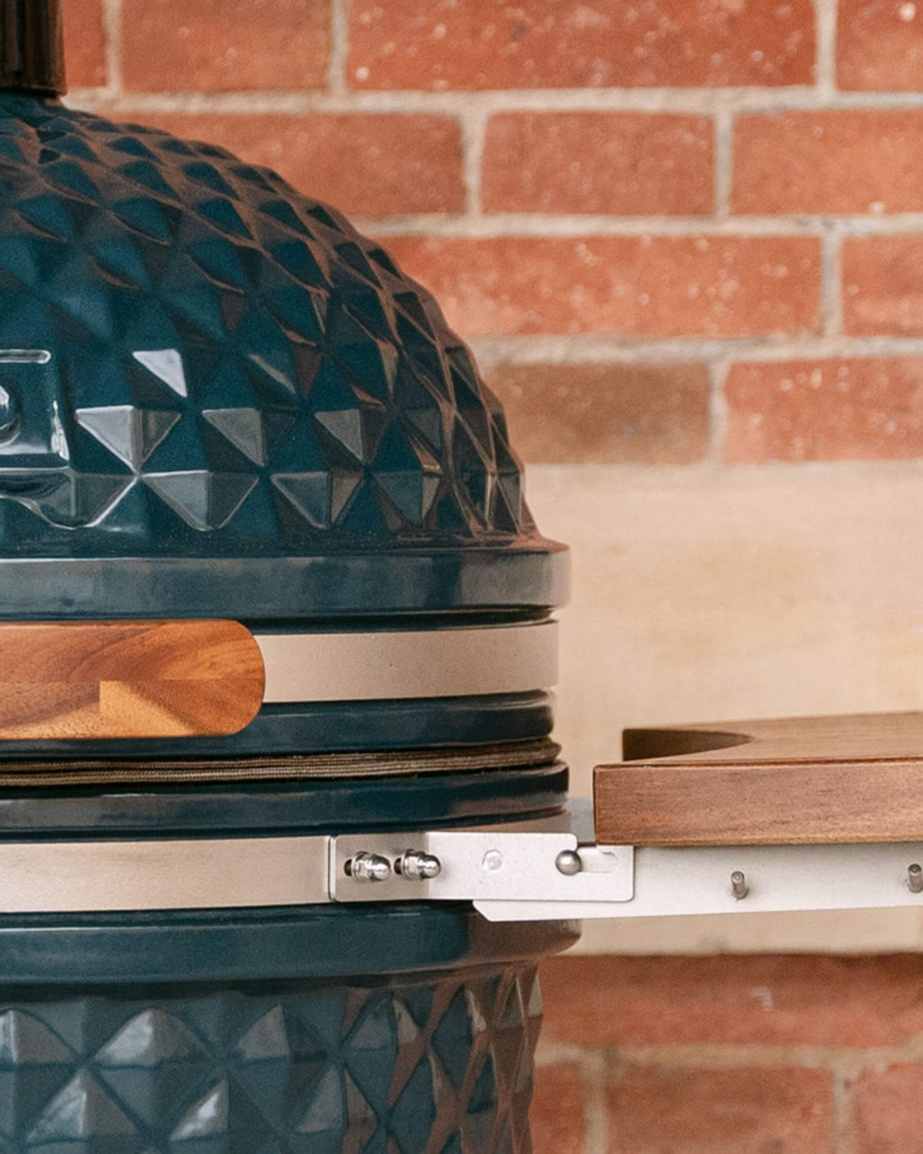 Detalhe das fixações metálicas que unem um tampo lateral em madeira ao corpo do kamado, com parafusos visíveis e vedante aparente.