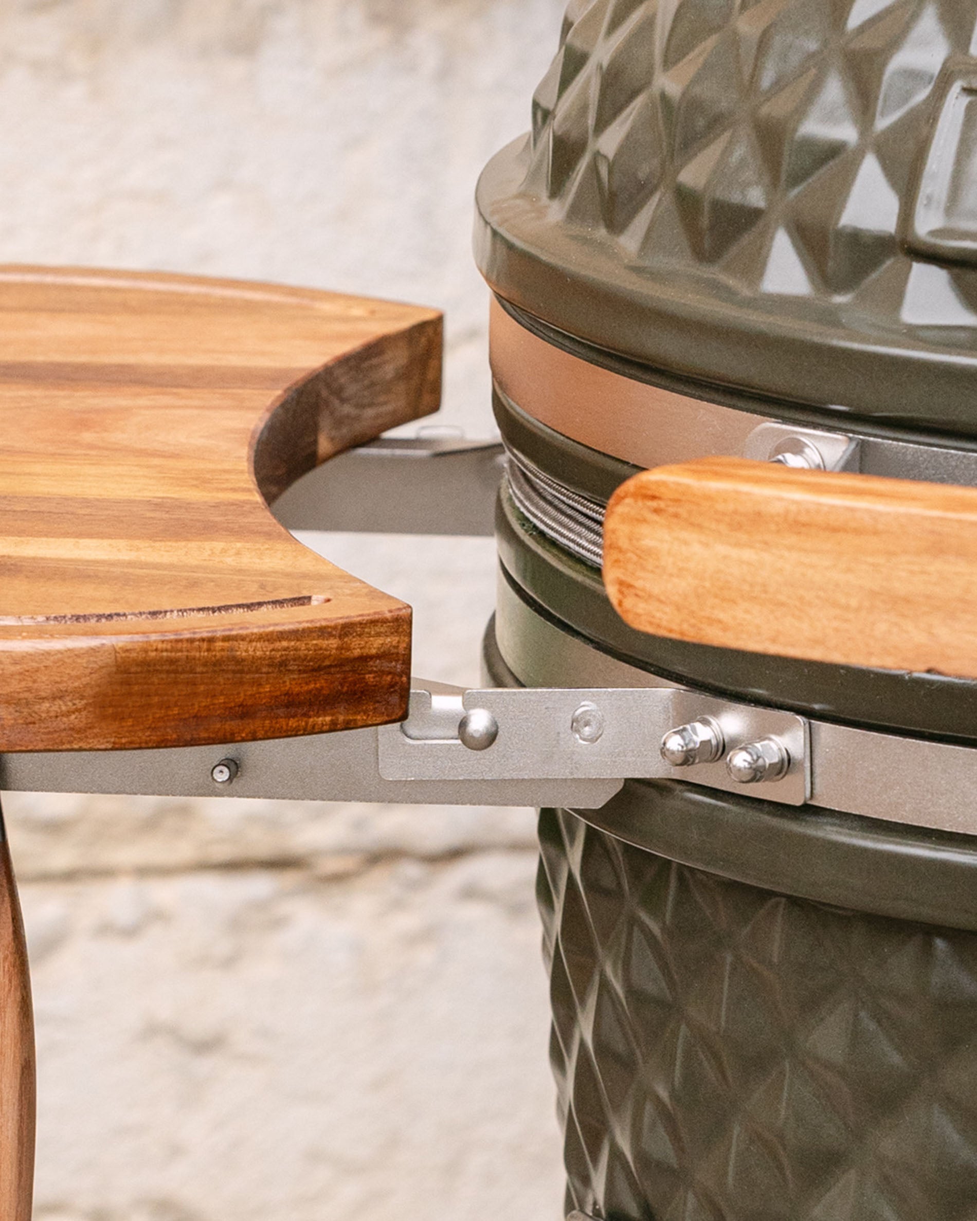 Detalhe das fixações metálicas que unem o tampo lateral em madeira ao kamado, com pega em madeira e aro em inox visíveis.
