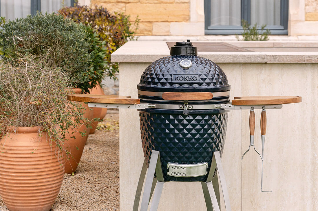Kamado Kokko instalado no exterior, com cerâmica preta texturada, prateleiras laterais em madeira, utensílios pendurados e estrutura metálica, integrado num espaço de cozinha de jardim.