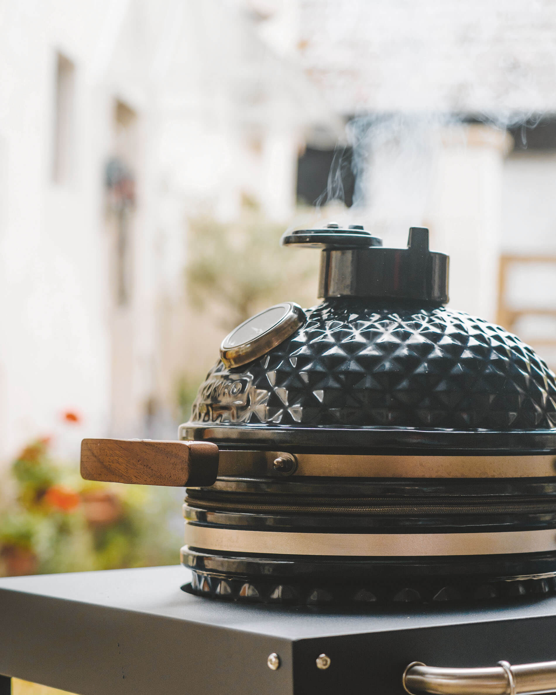 Kamado Kokko preto sobre carrinho, com tampa em cerâmica texturizada, pega em madeira, termómetro integrado e fumo a sair da chaminé.