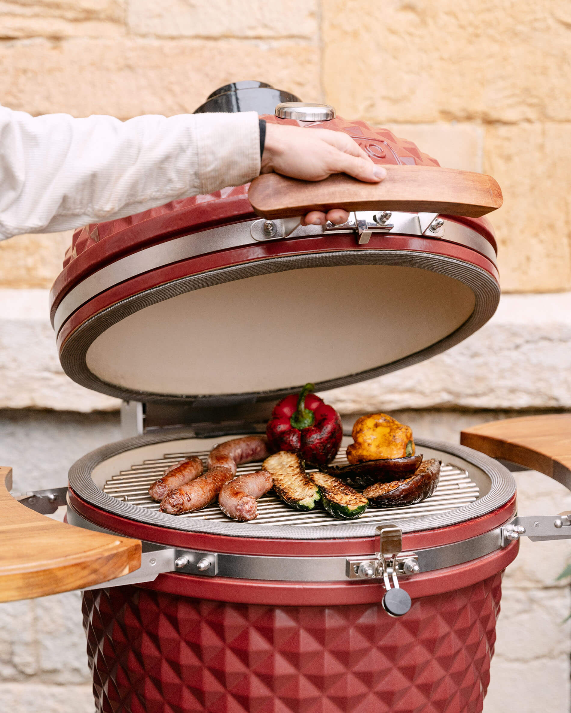 Kamado Kokko 22 polegadas em cerâmica vermelho óxido aberto, com salsichas e legumes a grelhar na grelha.